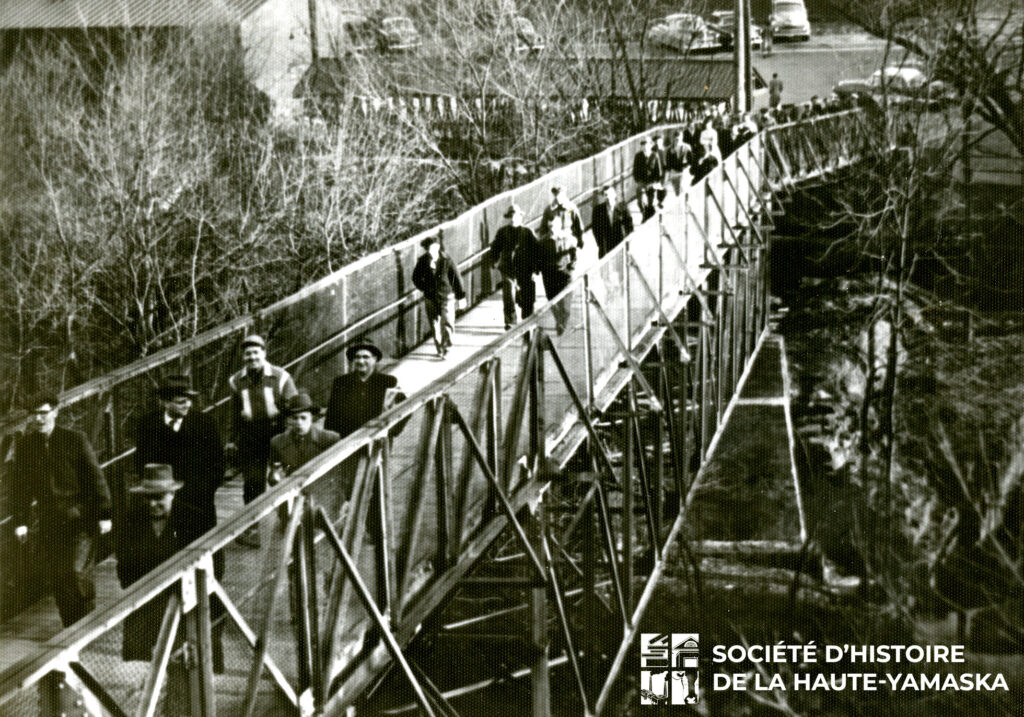 Une trentaine de travailleurs déambule sur la passerelle Miner. En arrière plan, deux bâtiments et une partie du stationnement de l'usine Miner Rubber. 